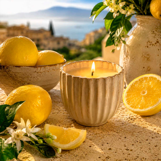 Candle in a textured container with lemons and flowers on a stone surface, with a scenic background.