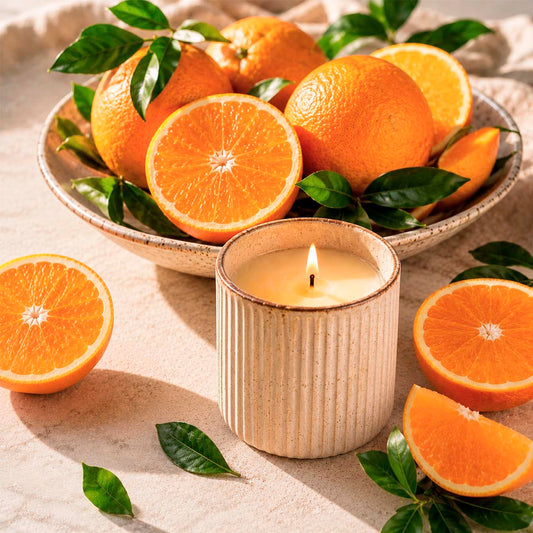 Burning candle in ceramic jar with fresh ripe oranges, orange slices and green citrus leaves in warm sunlight